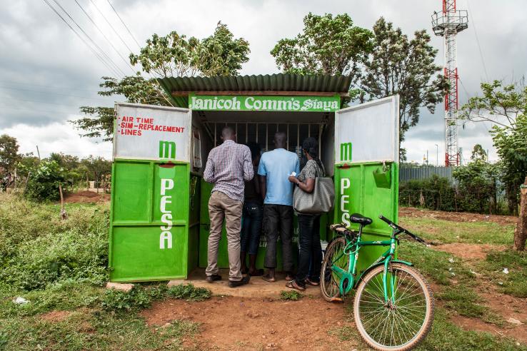 Three people stand at corrugated tin stand advertising mobile services in a field in Kenya