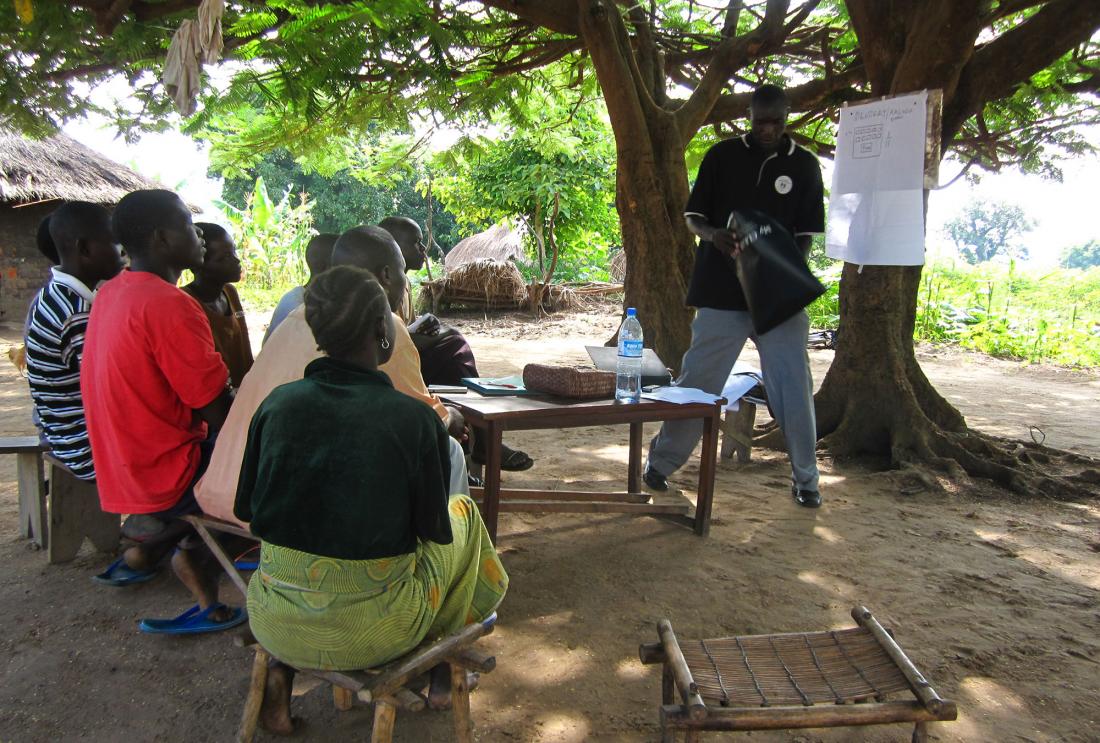 Group of Ugandan youth watch man giving presentation under a tree