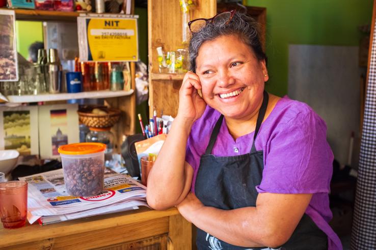 Image of a cheerful woman, wearing a purple shirt and an apron and smiling in a coffee shop setting.