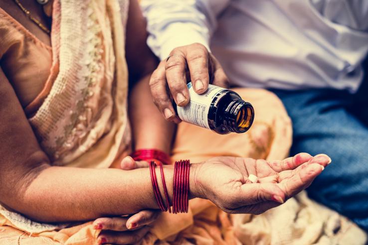 The photo depicts a close-up shot of a man distributing medication into the hands of a woman wearing a sari.