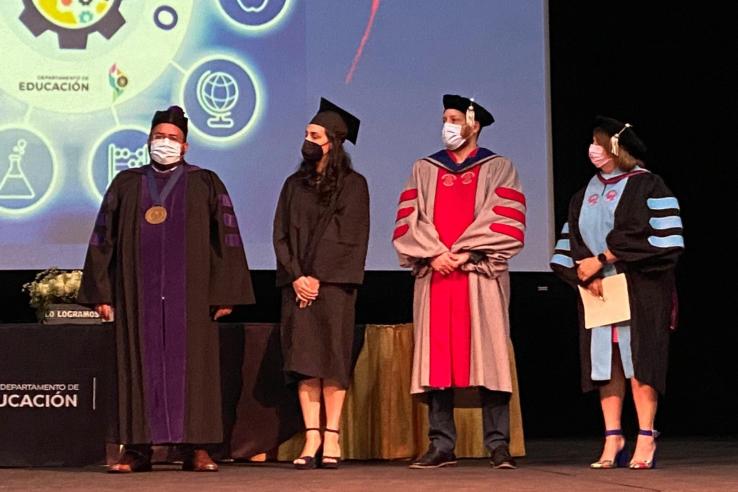 Four individuals in graduation robes stand on stage