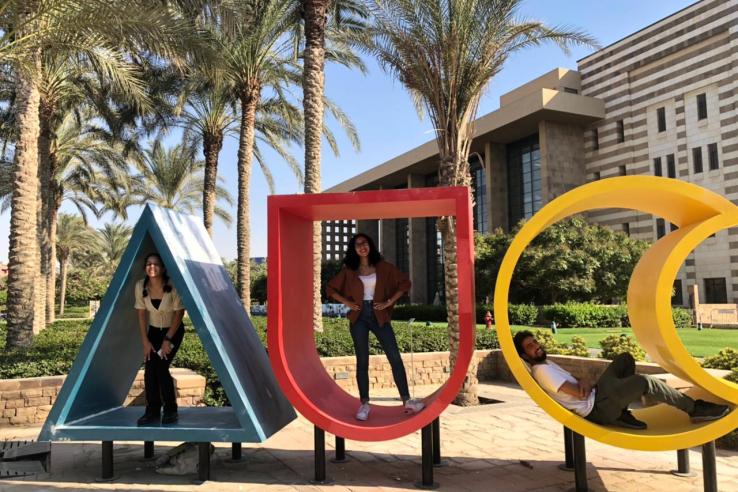 Three structures spell out "AUC" with palm trees in the background. One person sits inside each letter.