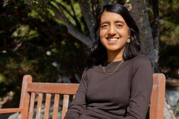 Seema Jayachandrani sits on a wooden bench outside and smiles at the camera.